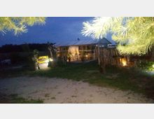 Ein beleuchtetes Gewächshaus mit Glaswänden in einem Garten bei Dämmerung, umgeben von Bäumen und begrenzt durch einen Holzzaun; im Vordergrund steht ein gelbes Schild mit der Aufschrift "Karmann".