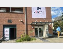 Backsteinfassade eines Gebäudes mit zwei Eingängen, einem Werbeschild „Conte’s Pizza & Pasta“ und einer großen Eiswaffel-Dekoration rechts. Neben dem Eingang stehen Pflanzen, ein Fahrrad sowie eine Bank, und auf der linken Seite ist ein weiteres Werbeschild für Abhol- und Lieferservice angebracht.