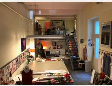 Offenes Atelier mit hoher Decke und Zwischengeschoss. Im Vordergrund ein großer Arbeitstisch mit Stoffen und Nähutensilien, umgeben von an der Wand befestigten Inspirationstafeln und Kleiderständern. Eine Treppe führt zu einer Galerie mit Regalen und Dekorationen.