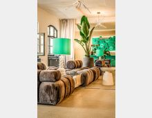 Modern gestaltete Lounge mit modularen, braunen Samtsofas, niedrigen Beistelltischen und großen grünen Tischlampen. Im Hintergrund stehen hohe Topfpflanzen vor einer grünen Barwand, dazu hohe Decken mit Sternleuchten und hohe Fenster mit Vorhängen.