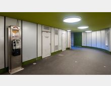 Ein unterirdischer Flur mit hellgrüner Decke, runden Deckenleuchten und weißen Wandpaneelen. Links steht eine Telefonzelle, während rechts Türen und ein grünes Gittertor zu sehen sind.