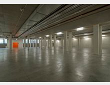 Großer, leerer Kellerraum mit glatten Betondecken, -wänden und -böden. Der Raum ist durch mehrere Stützpfeiler unterteilt, mit sichtbaren Lüftungsrohren und Kabelkanälen an der Decke; im Hintergrund befindet sich eine orangefarbene Tür und Fenster mit Tageslichteinfall.