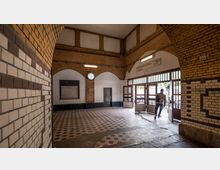 Innenansicht eines historischen Bahnhofsgebäudes mit gelblichen Klinkerwänden und gemusterten Fliesen auf dem Boden. Über dem Eingang hängt eine analoge Uhr, und durch die Glastüren ist ein Fahrradstellplatz im Freien sichtbar.