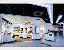 Moderner Verkaufsraum mit weißer Einrichtung und offenen Regalen, auf denen Kleidung, Accessoires und Merchandise-Artikel präsentiert werden. Auffällig sind geometrische Leuchten an der Decke, dunkler Fußboden und eine schlichte Gestaltung mit akzentuierten Beleuchtungselementen.