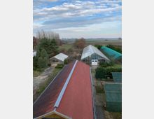 Blick auf eine ländliche Anlage mit Gewächshäusern, einem Gebäude mit rotem Ziegeldach im Vordergrund und Feldern im Hintergrund. Der Himmel ist blau mit vereinzelten Wolken.