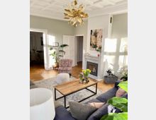 Wohnzimmer mit eleganter Einrichtung: ein Sofa mit Kissen, ein Couchtisch aus Holz, eine kunstvolle goldene Hängeleuchte, ein Kamin mit Marmorumrandung und darüber ein abstraktes Gemälde. Der Raum hat Parkettboden, helle Wände, hohe Decken mit Stuckelementen und wird von Tageslicht durchflutet.