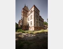 Historisches Backsteingebäude mit Türmen und verzierten Fassaden, umgeben von überwuchertem Gelände und einem gepflasterten Vorplatz; blauem Himmel und vereinzelten Bäumen im Hintergrund.