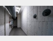 Ein schmaler, leerer Kellerflur mit sichtbaren Betonwänden und einer Metallbelüftungsrohrleitung an der Decke. An der linken Wand sind Elektrokästen und Kabel zu sehen, an der rechten Wand sind Wandhalterungen montiert.