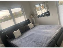 Schlafzimmer mit großem dunklem Polsterbett und grauer Tagesdecke, davor mehrere Kissen. An der Wand hängen ein Spiegel mit runden Lichtern, daneben stehen hohe Dekovasen; große Fenster mit Plissees lassen Tageslicht herein und geben den Blick auf Bäume frei.