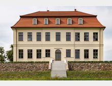 Historisches Gebäude mit cremefarbener Fassade und rotem Ziegeldach, symmetrisch gestalteten Fenstern und einem zentralen Eingang mit einer hohen Treppe. Vor dem Gebäude befindet sich eine Natursteinmauer mit einer niedrigen Hecke.