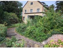 Zweistöckiges Haus mit hellgelber Fassade, umgeben von dichter Vegetation und einem kleinen Weg, der von einem bepflanzten Steingarten gesäumt ist. Im Hintergrund sind große Bäume zu sehen.