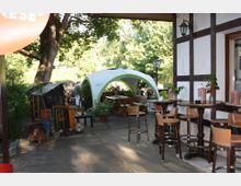Außenbereich eines Cafés oder Restaurants mit Holztischen und Stühlen, teilweise unter einem weißen Pavillon. Im Hintergrund stehen Bäume, während an der Fachwerkfassade links kleine Tische mit Dekoration sichtbar sind.