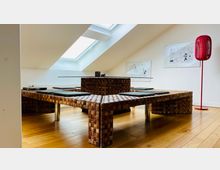 Modernes Zimmer mit minimalistischem Design, geprägt von einem niedrigen Tisch aus geflochtenem Holz mit Sitzkissen auf einem hellen Holzboden. Ein Dachfenster bringt Tageslicht herein, während an den weißen Wänden zwei gerahmte Bilder und eine rote Stehleuchte zu sehen sind.