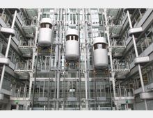 Moderner Innenhof eines Gebäudes mit drei verspiegelten Glasaufzügen in der Mitte, die an Schienen befestigt sind. Umgeben sind die Aufzüge von Glasfassaden, Balkonen und Stahlträgern in einer symmetrischen Anordnung.