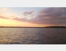 Sonnenuntergang über einem See mit leicht bewölktem Himmel. Im Vordergrund ist ruhiges Wasser, während am Horizont kleine Segelboote und eine bewaldete Uferlinie sichtbar sind.