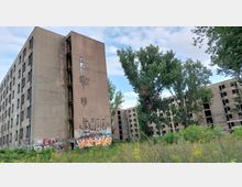 Eine verlassene, mehrstöckige Betonruine mit zahlreichen zerschlagenen Fenstern und Graffitis an den Fassaden. Im Vordergrund wachsen Gras und Bäume, die Umgebung wirkt wild und ungepflegt.