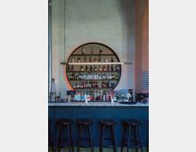 Eine moderne Bar mit einer halbkreisförmigen Regalwand im Hintergrund, die mit verschiedenen Spirituosenflaschen gefüllt ist. Vor der mit blauen Holzlatten verkleideten Theke stehen schwarze Hocker, und eine Hängelampe beleuchtet den Bereich darüber.