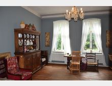 Wohnzimmer im historischen Stil mit blauen Wänden, einem Kronleuchter und Holzboden. Ein hölzernes Buffet mit dekorativem Porzellan, ein Esstisch mit Stühlen sowie zwei großen Fenstern mit weißen Vorhängen und einem Ausblick ins Grüne prägen den Raum.