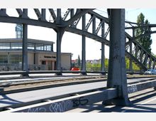 Eine Brückenkonstruktion aus grauem Metall mit sichtbaren Verstrebungen überspannt eine Straße und Straßenbahngleise. Im Hintergrund ist ein helles Gebäude mit großen Fenstern und einem Schild mit der Aufschrift „S Bornholmer Straße“ erkennbar, umgeben von Fahrrädern und Bäumen.