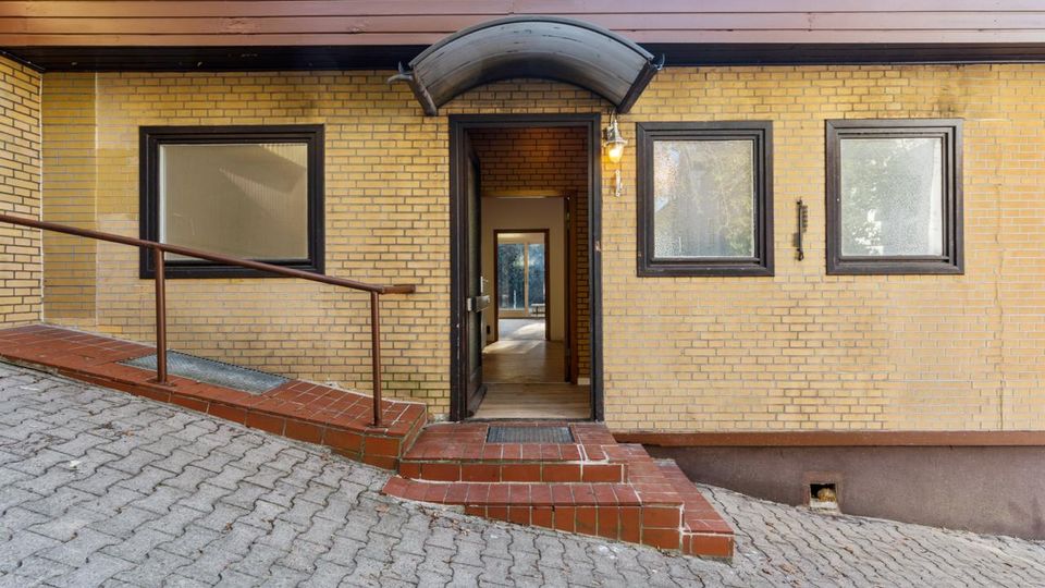 Eingangsbereich eines Backsteingebäudes mit offener Tür, kleinem gewölbtem Vordach und drei dunkel gerahmten Fenstern. Vor dem Eingang führen eine flache Rampe mit Geländer und niedrige Stufen über einen gepflasterten, leicht abfallenden Hof.