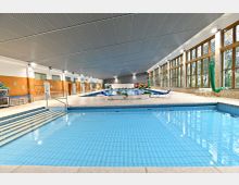 Innenraum eines Schwimmbads mit einem großen, flach abfallenden Becken im Vordergrund, einer zweiten Schwimmbahn im Hintergrund und einer breiten Fensterfront auf der rechten Seite. Der Raum hat helle Fliesen, orangefarbene Wandfliesen, Pflanzen zur Dekoration und Liegestühle entlang des Beckens.