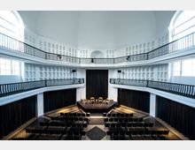 Innenansicht eines modernen, weißen Saals mit hoher Kuppeldecke. Der Raum verfügt über zwei Ebenen mit schwarzen Geländern, einen kleinen Bühnenbereich mit Holzstühlen und eine Bestuhlung aus Reihen schwarzer Stühle; seitlich sind große Fenster und schwarze Vorhänge sichtbar.