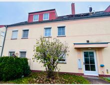 Mehrstöckiges Reihenhaus mit hellbeiger Fassade und rosa Akzenten, weißen Fenstern sowie rotem Ziegeldach mit Gaube, Schornstein und Satellitenschüssel. Vor dem Eingang mit kleinem Vordach stehen ein Strauch und eine Hecke in einem kleinen Vorgarten.