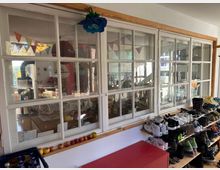 Ein heller Flur mit großen weißen Sprossenfenstern, die einen Blick in einen bunt geschmückten Innenraum mit Wimpelgirlanden und Möbeln ermöglichen. Entlang der Fenster steht ein offenes Schuhregal mit zahlreichen Schuhen, daneben liegt eine Reihe von Äpfeln auf einer Fensterbank.