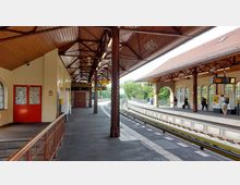 Bahnhof mit zwei überdachten Bahnsteigen, gestaltet in einem historischen Stil mit Holzdach und Metallträgern. Im Hintergrund sind Bäume sichtbar, und auf den Bahnsteigen befinden sich Fahrgäste sowie Fahrkartenautomaten und Informationsanzeigen.