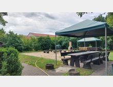 Ein Außenbereich mit Sitzplätzen unter zwei Pavillons, Holzbänken und -tischen. Im Hintergrund ist ein Grillplatz mit einem gemauerten Kamin und umliegenden Bäumen sichtbar.