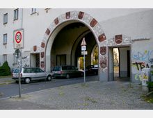 Ein historischer Torbogen mit dekorativen Verzierungen und Wappensymbolen, der einen Durchgang für Autos und Fußgänger bietet. Im Vordergrund sind geparkte Fahrzeuge, Straßenschilder und eine Kopfsteinpflasterfläche zu sehen, die Fassade ist mit Graffiti bemalt.