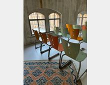 Reihe von bunten Holzstühlen auf grauem Boden in einem Raum mit großen, bogenförmigen Fenstern, durch die ein ziegelsteinernes Gebäude zu sehen ist; im Vordergrund ein Teppich mit geometrischem Muster.