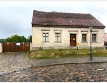 Ein altes, einstöckiges Wohnhaus mit verputzter Fassade, sichtbaren Abnutzungsspuren und einem roten Ziegeldach. Im Vordergrund befindet sich eine Kopfsteinpflasterstraße, eine Straßenlaterne und ein kleiner, ungepflegter Vorgarten mit niedriger Abgrenzung; links daneben ein Holztor vor einer Grundstückseinfahrt.