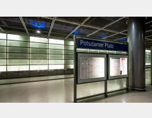 Unterirdischer Bahnhof mit einer beleuchteten Glaswand im Hintergrund. Im Vordergrund steht ein Schild mit der Aufschrift „Potsdamer Platz“ sowie Infotafeln darunter.