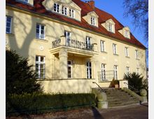 Gelbes zweistöckiges Gebäude im klassischen Stil mit rotem Satteldach, zahlreichen weißen Sprossenfenstern und einem kleinen Balkon mit schmiedeeisernem Geländer. Vor dem Eingang befinden sich eine Treppe mit steinernen Kugeln an den Seiten und umgebendes Grün mit Sträuchern.