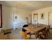 Ein einfach eingerichteter Raum mit Holzboden, einem großen Holztisch und verschiedenen Stühlen. An den weißen Wänden befinden sich ein kleines Wandregal und ein gerahmtes Bild, während im Hintergrund Türen und eine Holztreppe zu sehen sind.