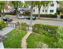 Ein gepflegter Vorgarten mit Rasen, einem schmalen Steinweg und einer Begrenzung durch eine Hecke und einen Metallzaun. Dahinter befindet sich eine gepflasterte Straße mit parkenden Autos und Wohnhäusern mit verschiedenen Fassaden sowie einzelnen Bäumen entlang der Straße.