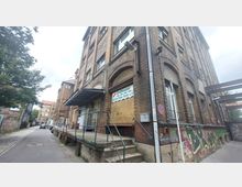 Mehrstöckiges Fabrikgebäude aus Backstein mit großen Rundbogenfenstern, einer überdachten Rampe und Graffiti an der Wand. Das Gebäude befindet sich an einer schmalen Straße mit weiteren historischen Gebäuden im Hintergrund.