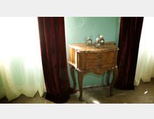 Ein kleiner, antiker Holzschrank mit geschwungenen Beinen und zwei Schubladen steht vor einer mintgrünen Wand zwischen zwei burgunderroten Vorhängen. Auf der Schrankoberfläche stehen drei dekorative Fläschchen mit goldenen Details.