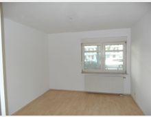 Leerstehender Raum mit weißen Wänden und hellem Laminatboden, einem großen Fenster mit Blick auf eine Straße und einem Heizkörper unter dem Fenster.