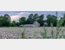 Eine ländliche Szene mit einem abgeernteten Feld im Vordergrund, dahinter ein altes Backsteingebäude mit einem roten Ziegeldach. Im Hintergrund stehen dichte Bäume und ein weiteres Gebäude, der Himmel ist bewölkt mit vereinzeltem Sonnenlicht.