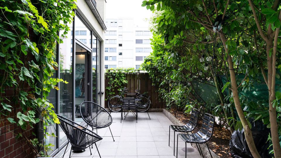Small urban patio beside a modern building with large glass doors and light gray tile flooring. The narrow courtyard is enclosed by dense trees and climbing vines, with black metal chairs and a small table, bicycles near a wooden fence, and string lights hanging in the greenery.