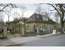 Kleines Gebäude im klassischen Stil mit Satteldach und grünen Fensterläden, gelegen hinter einem schmiedeeisernen Tor mit seitlichen Laternen. Im Vordergrund ist eine gepflasterte Straße, während im Hintergrund blattlose Bäume und eine Gartenanlage zu sehen sind.