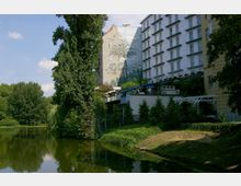 Ein kleiner See mit grüner Vegetation und Bäumen im Vordergrund, daneben mehrstöckige Wohn- und Geschäftsgebäude, von denen eines eine gemalte Fassade mit Palmenmotiven zeigt. Im unteren Bereich der Gebäude befinden sich Terrassen mit Überdachungen und Pflanzen.