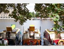 Drei kleine, farbige Hausboote sind nebeneinander an einem Holzsteg auf einem Fluss festgemacht, im Vordergrund rahmen Baumzweige die Szene ein. Im Hintergrund stehen Gebäude und Bäume entlang des gegenüberliegenden Ufers.