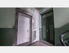 Treppenhaus eines älteren Gebäudes mit einer weißen Tür links und einer dunkelgrünen Tür mit Glaseinsätzen rechts, umgeben von Wänden mit grüner Farbe und teils abgeblättertem Putz. Der Boden ist mit einem gemusterten Teppich oder Belag ausgestattet, und ein schwarzes Geländer ist am rechten Bildrand sichtbar.