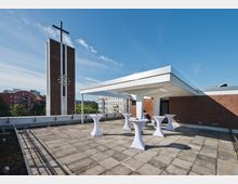 Dachterrasse mit Steinplattenboden, weißen Stehtischen und einer Überdachung auf einer Seite; im Hintergrund ein roter Kirchturm mit Kreuz und Uhr sowie umliegende Gebäude.