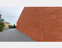 Eine lange, geschwungene Ziegelsteinwand aus rotem Backstein verläuft entlang eines gepflasterten Gehwegs. Im Hintergrund sind Bäume, ein gelbes Wohnhaus und parkende Autos zu sehen.