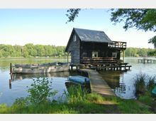 Holzhaus auf Stelzen am Ufer eines Sees, verbunden mit einem schmalen Holzsteg. Im Vordergrund liegt ein Ruderboot im Wasser, umgeben von Schilf und Bäumen, während im Hintergrund ein dichter Wald das Seeufer säumt.