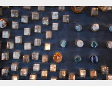 Nahaufnahme einer mit dunkelblauem Stoff bedeckten Fläche, auf der rechteckige und runde Glaselemente in verschiedenen Farben, überwiegend klar, einige bernsteinfarben oder grünlich, in einem regelmäßigen Muster angebracht sind.